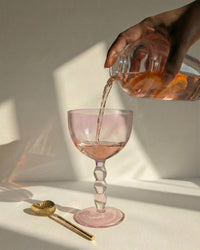 A hand pours a pink drink from a glass pitcher into an elegant, pink-stemmed glass. Sunlight casts soft shadows beside Casa Amarosa barware and Casa Amarosa Heart Shaped Brass Cocktail Spoons (Set of 4).