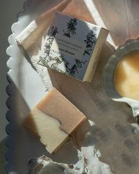 A bar of The Quiet Botanist’s Lavender Fields - Bar Soap in a floral box rests on a white cloth atop a tray, beside a dish with cream. Sunlight casts soft shadows, highlighting the handmade soap’s elegant, natural charm.