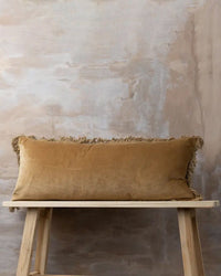 The Saarde Velvet Body Pillow w/ Feather Insert in Nutmeg rests on a wooden bench against a textured beige wall, its rich tan velvet and fringed edges adding warmth and elegance to the scene.