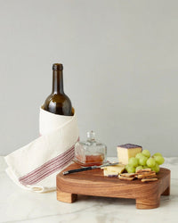 A wine bottle in a white cloth with red stripes stands behind the etúHOME New Classic Rectangle Trivet Riser, Small, which displays cheese, grapes, crackers, honey in a glass jar, and a knife on a marble surface—an elegant kitchen essential.