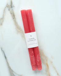 Two unscented Cherry Taper Candles by BOTANICA, wrapped with a label reading “Taper Candles,” rest on a light-colored marble surface.