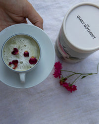 A hand holds a cup of green tea topped with Edible Rose Petals by The Quiet Botanist, resting on a white saucer. Beside it sits a branded canister and a small pink flower sprig on a white cloth.