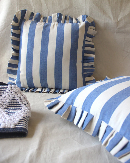 Two Casa Amarosa Denim Ruffle 16x16in pillows with blue and white stripes and ruffled edges rest on a light fabric surface, accompanied by a textured, folded blue-and-white patterned cloth nearby.