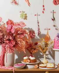 The Flora Nouveaux by Chronicle Books decorates a pastel table with pink and yellow floral arrangements, cakes, and tarts, while modern dried flowers accent the white wall for a bright, festive look.