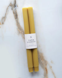 Two Martini Olive Fluted Taper Candles by BOTANICA with white labels lie side by side on a white marble surface with light brown veins, highlighting the elegance of hand-dyed candles.
