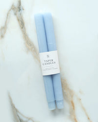Two hand-dyed Cornflower Blue Taper Candles by BOTANICA, wrapped in a white label, rest on a white marble surface with subtle gray and beige veining.