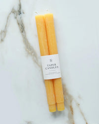 Two yellow Goldenrod Taper Candles by BOTANICA, each with a white "Taper Candles" label, are displayed side by side on a white marble surface with brownish-gray veining.