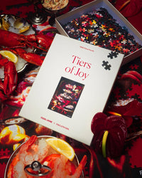 A Piecework Puzzles "Tiers of Joy - Food+Wine Collab 1000 Piece Puzzle" sits on a table, partially assembled, with lobster, shrimp on ice with lemon slices, and red decorative flowers surrounding it.