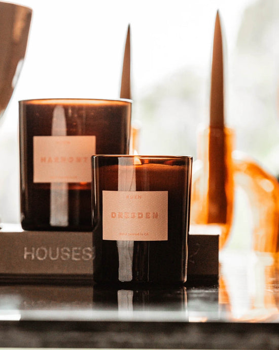 Two brown glass ROEN candles—one the vegan soy wax Dresden Candle, the other labeled Marigot—rest on a reflective surface with blurred decor and soft lighting in the background.