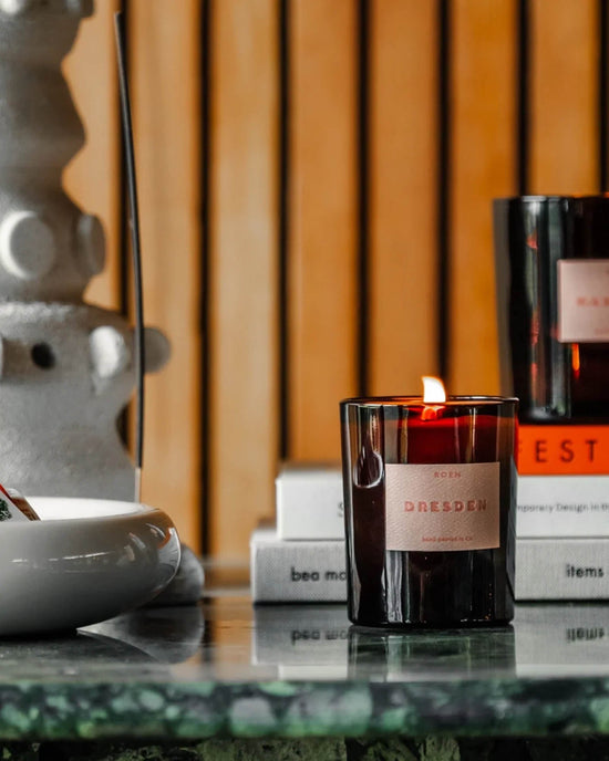 A lit Dresden Candle by ROEN—black glass, vegan soy wax—rests on marble beside a bowl and stacked books, with an unlit candle and abstract decor in the background.