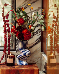A striped ceramic vase with red and white flowers sits on wood beside a lit ROEN Marmont Candle. The setting features marble accents and windows in the background.