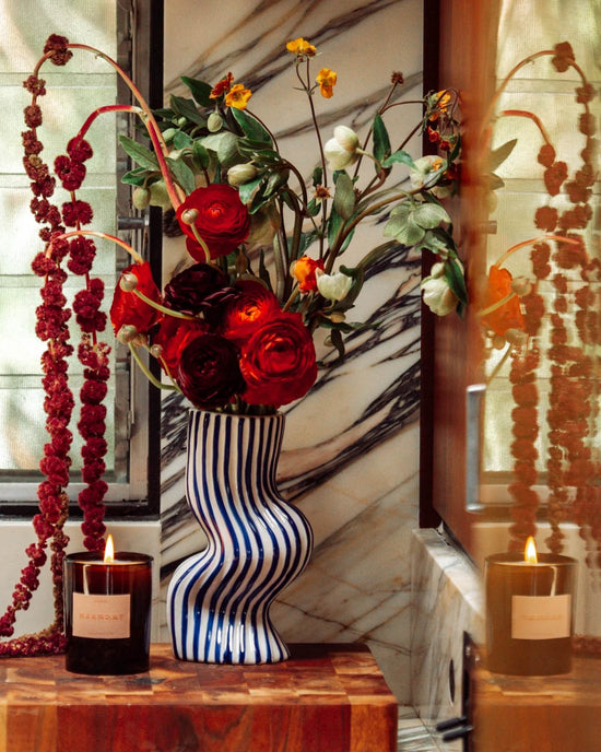 A striped ceramic vase with red and white flowers sits on wood beside a lit ROEN Marmont Candle. The setting features marble accents and windows in the background.