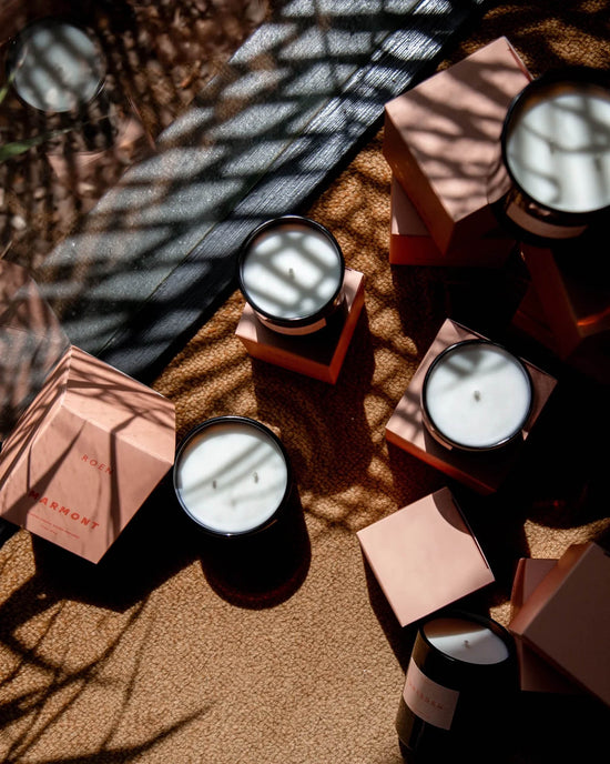 ROEN’s Marmont Candle—white coconut soy wax in black jars—sits on pink hexagonal boxes atop a tan surface with patterned shadows, elegantly evoking a Hollywood hotel fragrance.