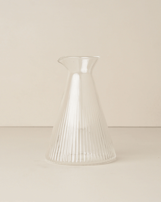 The Saarde Kairos Angular Carafe in Clear, featuring vertical ribbed lines, rests on a neutral surface against a light background, accentuating its elegant design.