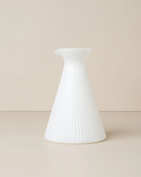 The Kairos Angular Carafe in Opaque White by Saarde features a cone-shaped ceramic form with a narrow spout and subtle vertical ridges, reminiscent of ribbed glass, showcased against a plain beige background.