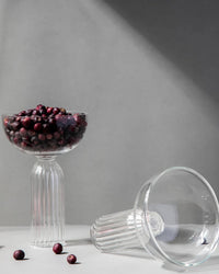 Two ribbed Cocktail Glasses by Casa Amarosa are displayed—one upright, filled with dark berries, the other lying empty on its side. Handcrafted in India, both rest on a sunlit surface with scattered berries against a gray backdrop.
