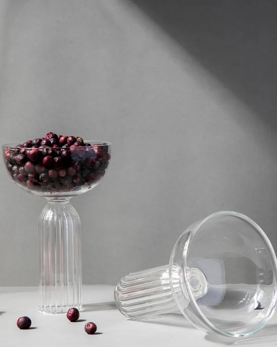 Two ribbed Cocktail Glasses by Casa Amarosa are displayed—one upright, filled with dark berries, the other lying empty on its side. Handcrafted in India, both rest on a sunlit surface with scattered berries against a gray backdrop.