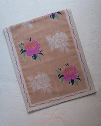 The Babita Table Runner by Soil to Studio is a rectangular linen runner in beige with pink and white floral patterns, green leaves, and a pink wavy border—part of the Valley of Flowers Collection.