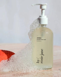 A clear pump bottle of Rosie Jane James Everyday Body Wash, a hydrating cleanser, is partially covered with soap bubbles on a light surface, with an orange textured object in the background.