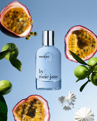 A bottle of Rosie Jane Eau De Parfum in Matilda sits among halved passion fruits, green limes on branches, and white meringues, set against a light blue background.