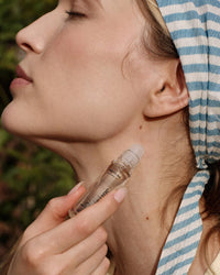 A woman in a blue-and-white striped headscarf applies Rosie Jane Perfume Oil in Matilda to her neck, eyes closed, surrounded by lush greenery.