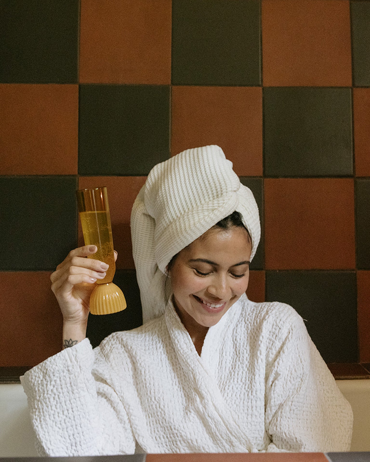 Woman in a white robe with a towel on her head, holding a glass of orange juice against a checkered wall.
