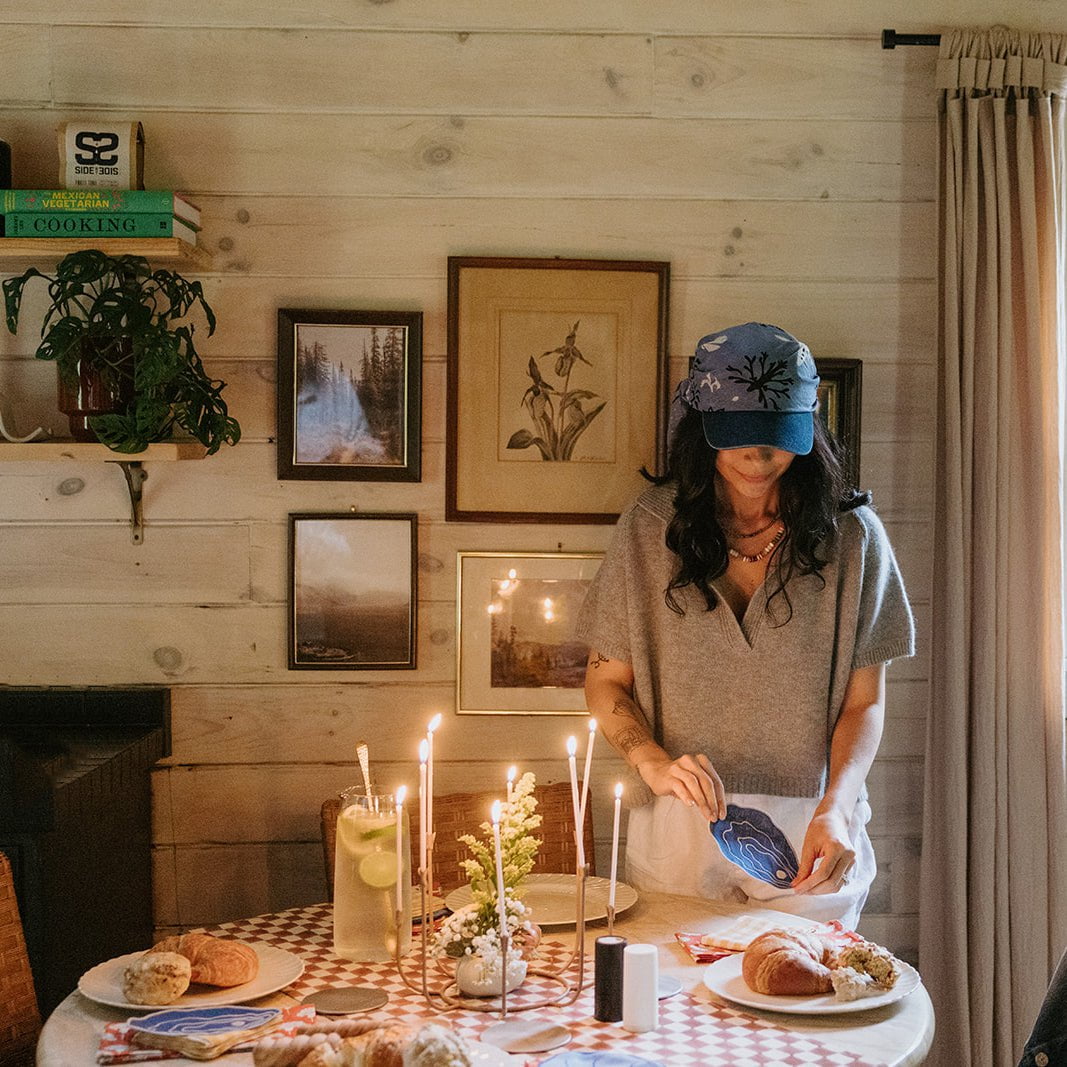 Woman setting a table with candles and food in a cozy room with wooden walls and framed pictures.