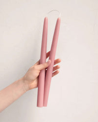 A hand displays a pair of 12in Beeswax Taper Candles by The Floral Society, connected and hand-dipped in pink, against a light textured background.