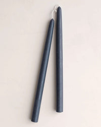 Two tall, dark blue 12in Beeswax Taper Candles from The Floral Society with white wicks hang side by side against a plain, light background.