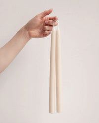 A hand holds two 12in Beeswax Taper Candles by The Floral Society by their wicks against a plain, light background.