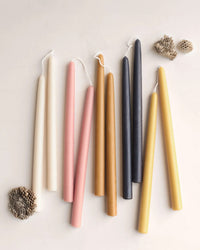 Eight 12in Beeswax Taper Candles by The Floral Society—hand-dipped in cream, pink, brown, black, and yellow—are arranged on a light surface next to two honeycomb pieces.