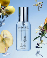 A clear bottle of Rosie Jane Birthday Suit Body + Hair Perfume Mist is centered on a blue backdrop, surrounded by apple slices, white flowers, green leaves, and small yellow fruit for a fresh, clean scent.