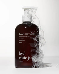 A dark bottle of Rosie Jane Calm Body Wash, featuring lavender, chamomile, and neroli on the label, stands against a light background with bubbles foaming along its side.