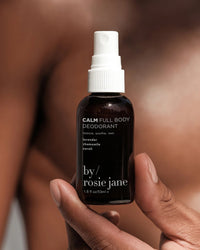 A hand holds a small dark spray bottle labeled Calm Full Body Deodorant by Rosie Jane—an aluminum-free deodorant with lavender, chamomile, and neroli. The person’s softly blurred bare chest is visible in the background.