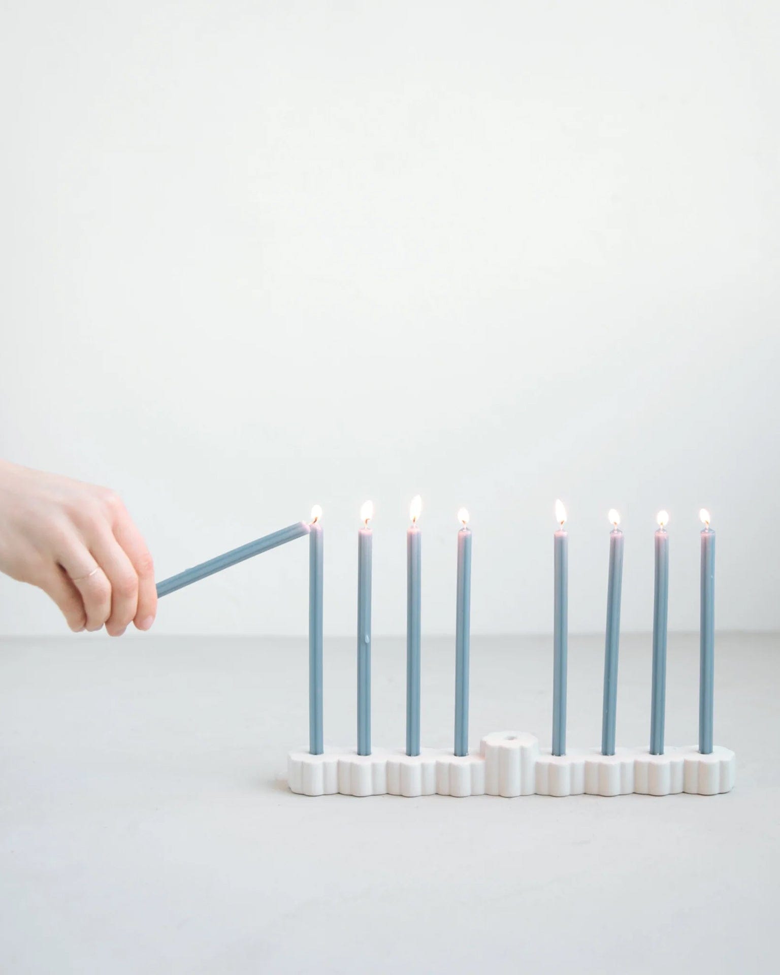 A hand lights one of nine Hanukkah candles in The Floral Society’s Fleur Menorah, a modern white ceramic piece, set on a light surface against a plain white background.