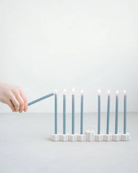 A hand lights one of nine Hanukkah candles in The Floral Society’s Fleur Menorah, a modern white ceramic piece, set on a light surface against a plain white background.