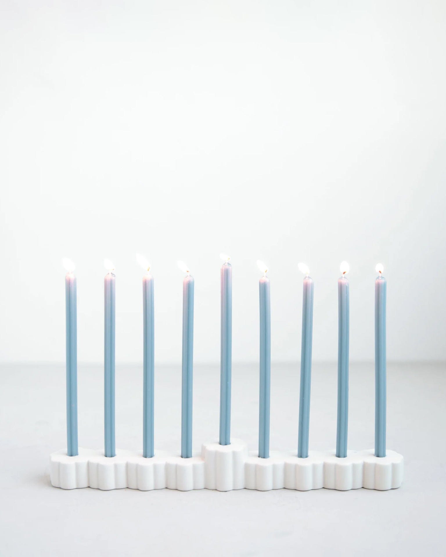 The Fleur Menorah by The Floral Society, a modern white ceramic piece, holds nine blue Hanukkah candles, each with a glowing flame against a plain light background.