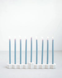 The Fleur Menorah by The Floral Society, a modern white ceramic piece, holds nine blue Hanukkah candles, each with a glowing flame against a plain light background.
