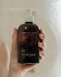 A hand holds a brown bottle of Chill Body Wash by Rosie Jane, featuring a white pump, seen through foggy glass. The label notes peppermint, mandarin, and cypress for an invigorating scent.