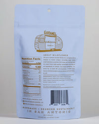 The back of a light blue Wildflower Caramel Co Coconut Caramels (Vegan) in A Bag shows nutrition facts, ingredients, a barcode, the company story, and details on handmade, sustainably sourced production in San Antonio.