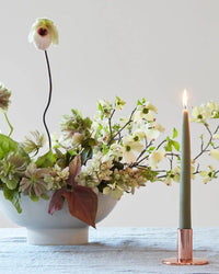 A white bowl with green and white flowers sits on a light blue tablecloth next to The Floral Society’s Copper Taper Holder - Medium with a lit green taper candle, all set against a plain light background.
