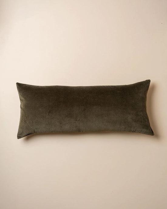 The Saarde Velvet Body Pillow w/ Feather Insert in Dark Olive rests on a plain beige surface.