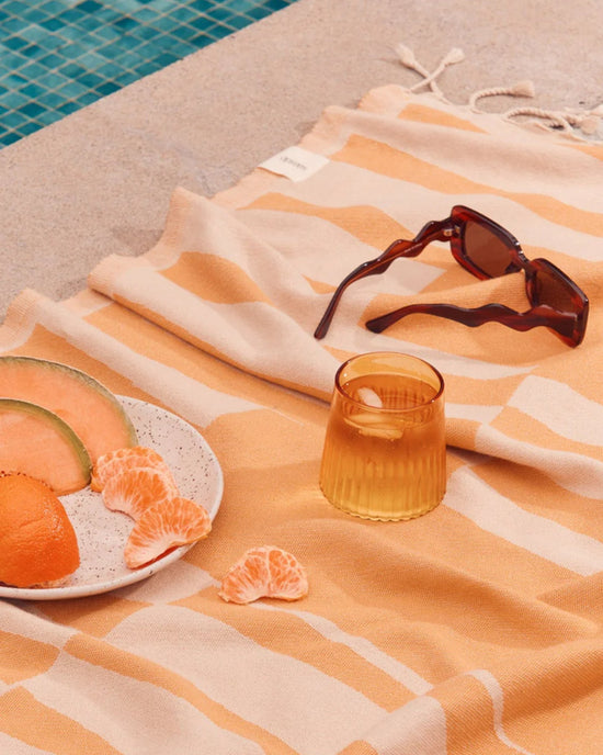 A patterned towel by the poolside is topped with sunglasses, a Saarde Kairos Medium Water Glass in Yellow Topaz, and a plate of sliced melon, orange, and tangerine.