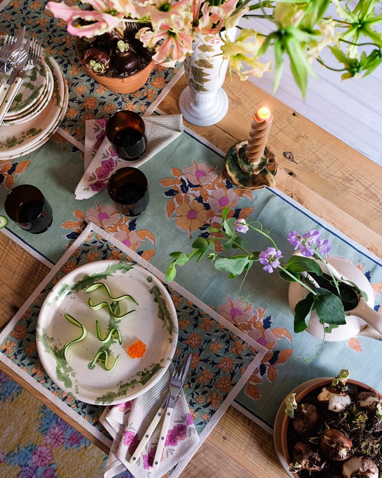 A wooden table set with plates, cutlery, glasses, a candle, flowers, and food is decorated with handmade linens and Soil to Studios Simran Table Napkins S/4 for a colorful and inviting scene.