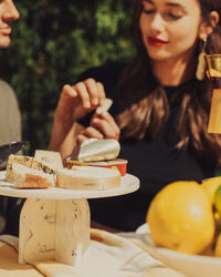 A woman with long brown hair and red lipstick sits at a table preparing food. In the foreground, the Saarde Flow Resin Cake Stand in Merle displays assorted cheeses atop a yellow tablecloth with fruit.