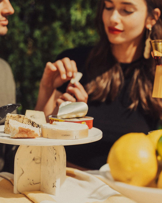 A woman with long brown hair and red lipstick sits at a table preparing food. In the foreground, the Saarde Flow Resin Cake Stand in Merle displays assorted cheeses atop a yellow tablecloth with fruit.