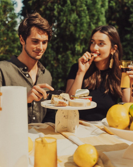 A young man and woman enjoy a sunny picnic outdoors with lemons, glasses, and the Saarde Flow Resin Cake Stand in Merle. Surrounded by greenery, he slices cheese while she eats, both relishing the handmade stand’s marble finish.