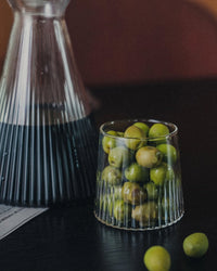 A Kairos Medium Water Glass in Clear by Saarde, filled with green olives, sits beside a ribbed carafe holding a dark liquid on a black table.