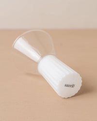 The Saarde Kairos Wine Glass in Opaque White, featuring a ribbed white base, rests on its side atop a beige surface.