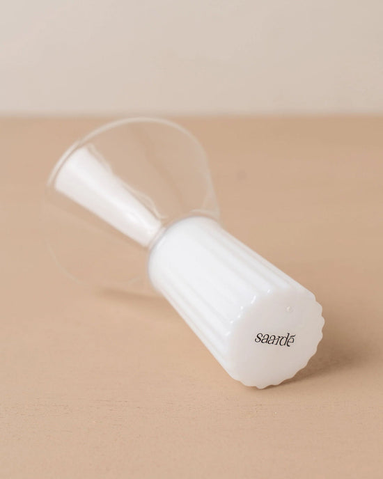 The Saarde Kairos Wine Glass in Opaque White, featuring a ribbed white base, rests on its side atop a beige surface.